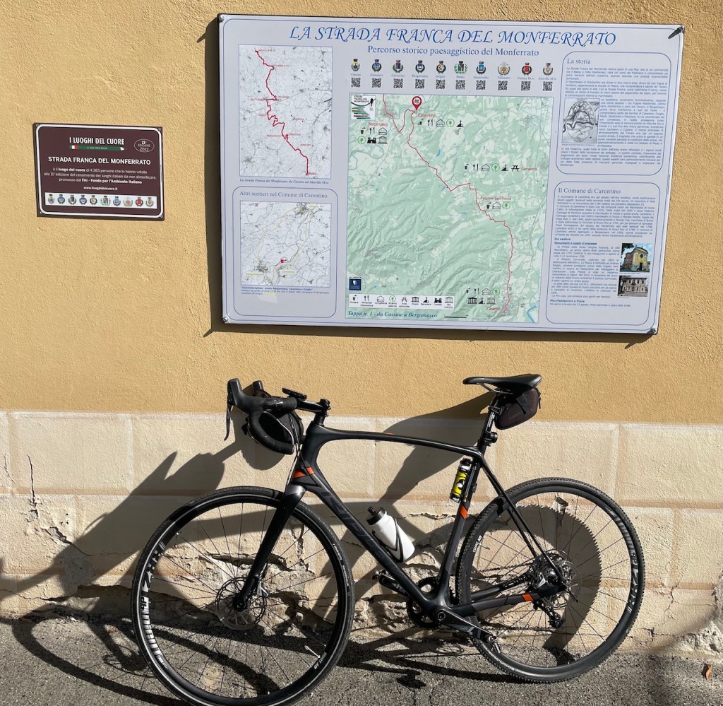 Bicicletta nera appoggiata a un muro con una mappa della Strada Franca del Monferrato e un pannello informativo accanto.