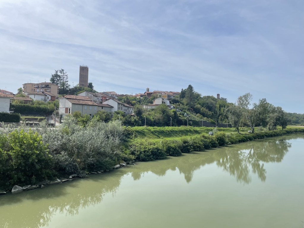 Panorama di un villaggio collinare con case e una torre, riflesso nel fiume di colore verde.